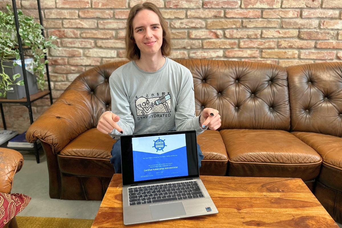 Eine Person sitzt auf einem braunen Ledersofa vor einer Ziegelwand und zeigt stolz auf einen Laptop, auf dem die bestandene Prüfung zum „Certified Kubernetes Administrator“ angezeigt wird – das Bild symbolisiert beruflichen Erfolg und IT-Expertise.